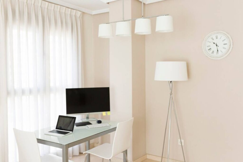 Modern home office with laptop on a sleek desk, large monitor, and stylish hanging lights.