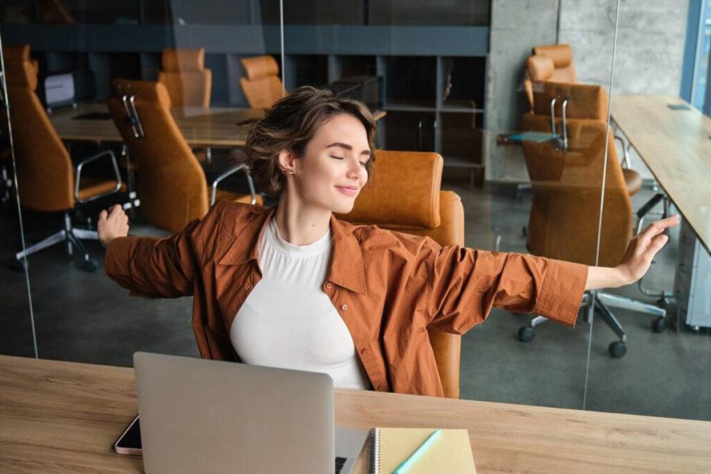 Woman stretching with closed eyes and arms outstretched, seated at a desk in a modern office space.