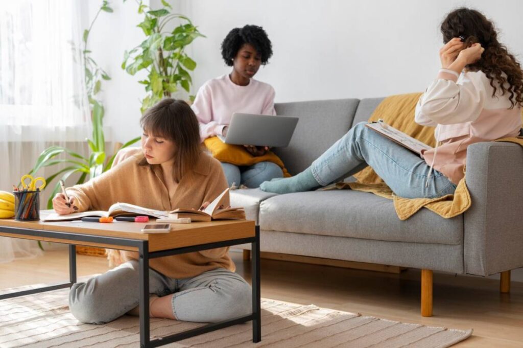 Three young women studying and working in a bright living room, one writing, another using a laptop