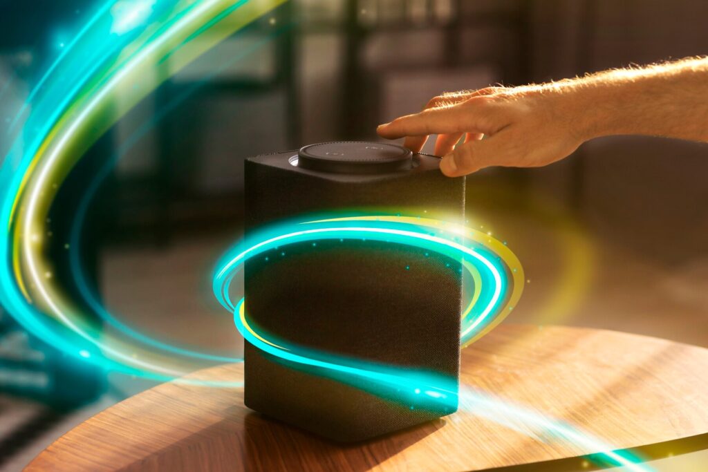 A hand adjusts a modern speaker on a wooden table, surrounded by colorful swirling light effects in a bright room.