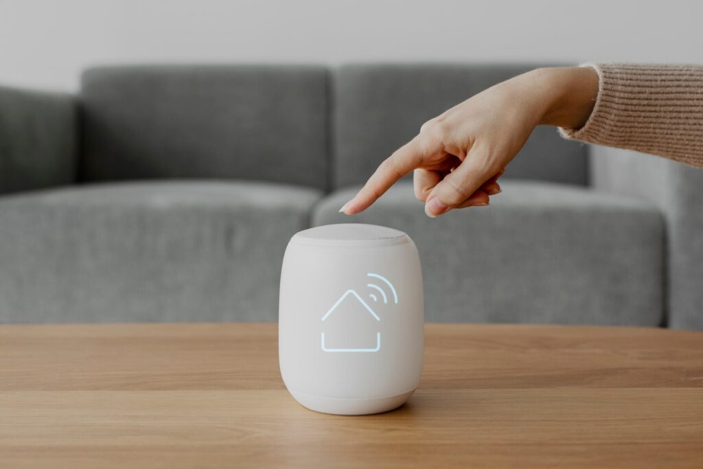 A hand presses a smart home device with a house icon, placed on a wooden table in a cozy, gray-furnished living room.