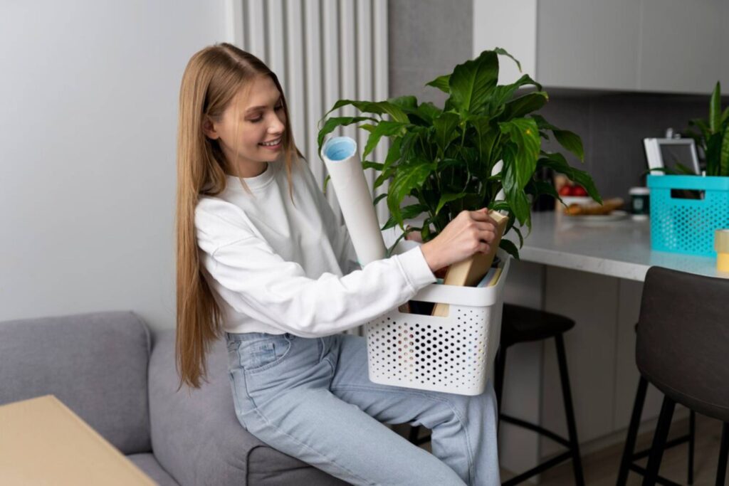 Young woman sits on a sofa, arranging a large potted plant inside a portable white bin in a modern kitchen setting.