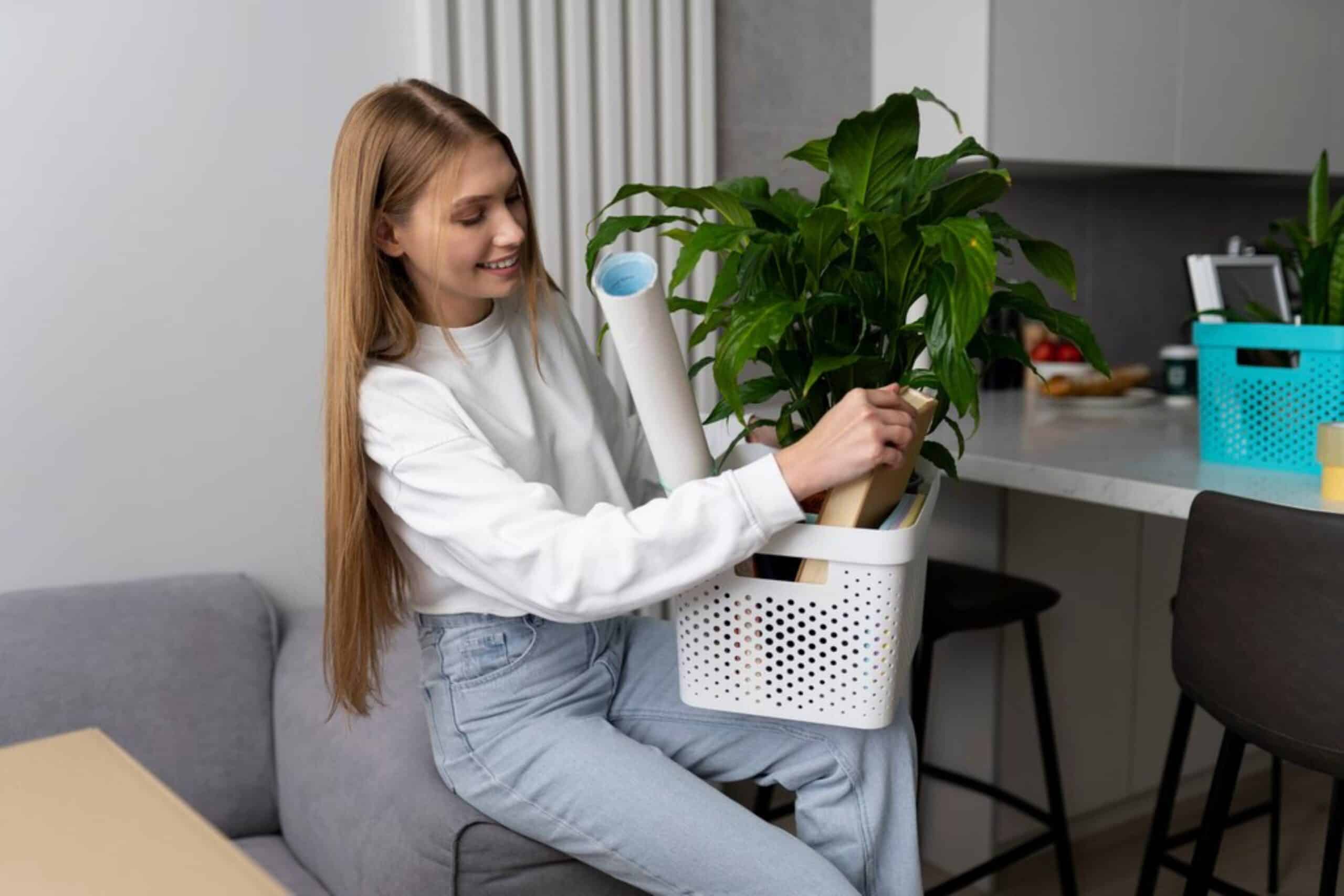 Young woman sits on a sofa, arranging a large potted plant inside a portable white bin in a modern kitchen setting.