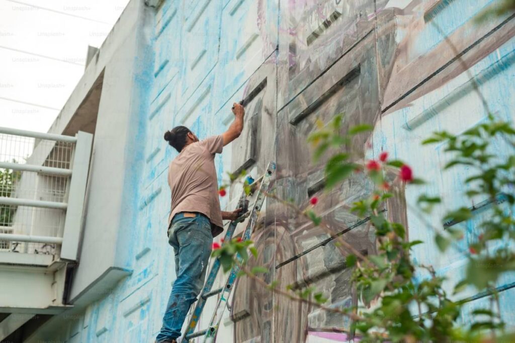 Man on ladder painting a large mural on an urban building wall.